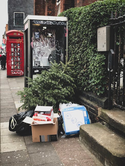 A collection of various discarded items, including a small pine tree, a cardboard box, a blue and white box, and a black bag, placed on the pavement near a neatly trimmed green hedge and a black metal fence. Behind the rubbish, there is a small glass-fronted kiosk or shelter with graffiti and stickers on its surface, and a red traditional British postal box positioned further to the left. The scene is set on a quiet urban street corner with brick buildings visible in the background, and the pavement appears clean aside from the waste. The placement of the waste suggests it has been temporarily left for collection or clearance. The greenery and urban environment are typical of a residential or commercial area in Merton, and the presence of the Rubbish Removal Merton service is implied through the context of waste management activity.