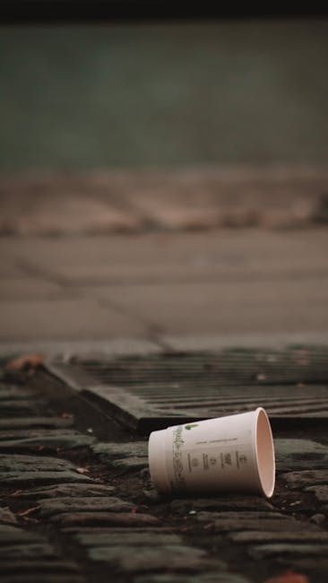 A white paper coffee cup with a plastic lid lies tipped over on a weathered wooden surface, near a metal grate embedded in the ground. The area appears to be an outdoor pavement or pathway with dark, uneven tiles and a slightly damp appearance. In the background, there is a blurred, dark surface which could be a street or driveway, with soft natural lighting suggesting an overcast day. The scene depicts an instance of litter possibly collected during an outdoor rubbish removal or waste clearance process, aligned with private waste handling services such as those offered by Rubbish Removal Merton. The focus on the discarded cup and the surrounding environment highlights the importance of proper rubbish disposal practices in urban or commercial settings, supporting the concept of independent collection or on-site rubbish clearance.