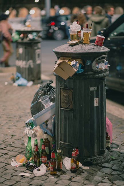 A collection of various discarded items, including a small pine tree, a cardboard box, a blue and white box, and a black bag, placed on the pavement near a neatly trimmed green hedge and a black metal fence. Behind the rubbish, there is a small glass-fronted kiosk or shelter with graffiti and stickers on its surface, and a red traditional British postal box positioned further to the left. The scene is set on a quiet urban street corner with brick buildings visible in the background, and the pavement appears clean aside from the waste. The placement of the waste suggests it has been temporarily left for collection or clearance. The greenery and urban environment are typical of a residential or commercial area in Merton, and the presence of the Rubbish Removal Merton service is implied through the context of waste management activity.