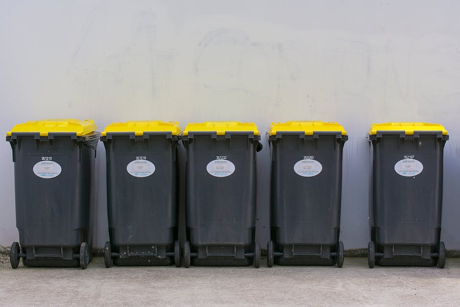 A black cylindrical waste bin with a domed lid, located on a cobblestone street, is visibly overflowing with various types of rubbish. On top of the bin, there are disposable cups with plastic lids containing spilled drinks, a crumpled paper napkin, and a cardboard food container. Hanging from the side of the bin are additional rubbish items, including plastic bottles, paper packaging, and an empty food carton. Surrounding the base of the bin are discarded bottles, some tipped over with liquid inside, along with crumpled paper, food wrappers, and other small waste items scattered on the ground. In the background, a blurred scene of parked cars, pedestrians, and a city street suggests an outdoor social area or busy urban environment. The overall scene underscores the common issue of littering and the need for effective waste management solutions, such as private rubbish collection or on-site clearance, managed by services like Rubbish Removal Merton.