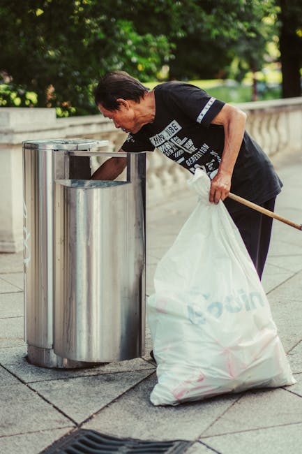 A man with dark hair, wearing a black t-shirt with white printed text, is seen standing next to a metal rubbish bin outdoors, in a location with a paved surface and a stone balustrade. He is leaning forward, holding a white garbage bag in his left hand, which appears to be filled with waste, and using his right hand to reach into the open-top container. The bin is cylindrical with a smooth, reflective stainless steel finish, positioned against a backdrop of greenery and trees, suggesting an urban park or promenade setting. The scene is well-lit with natural daylight, highlighting the textures of the stainless steel, the man's clothing, and the surrounding environment. This image exemplifies on-site rubbish collection and disposal, showing a private waste handling scenario consistent with independent rubbish removal services such as those provided by Rubbish Removal Merton, emphasizing proper waste management practices.