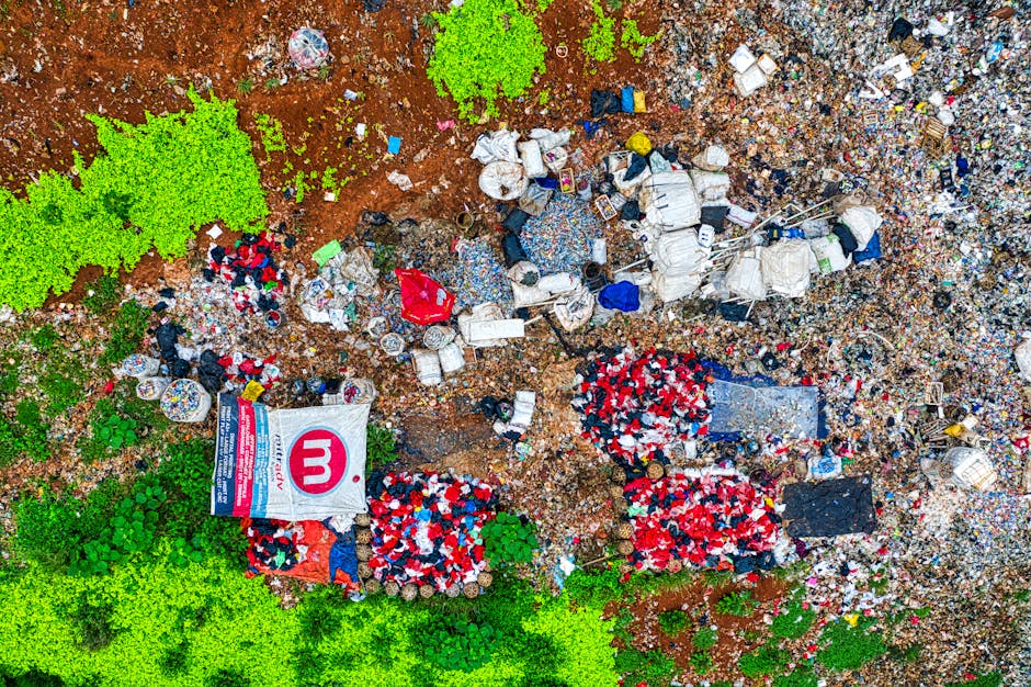 An aerial view of a large area of landfill or waste disposal site showing a mix of household and industrial waste, including many plastic bottles, paper, and packaging materials scattered across the ground. The site features several piles of discarded items, some covered with plastic sheeting or tarps, and a prominently visible bag with a large pink 'm' logo. The surrounding environment includes patches of green vegetation and exposed earth, with areas of brown soil and broken-up ground material. There are some small trees or bushes on the perimeter, indicating the site is outdoors. The waste appears to be a combination of mixed refuse, possibly from collection or disposal activities, with no visible machinery or equipment present. The scene reflects a typical waste accumulation area which relates to private rubbish handling or alternative collection methods, as managed by companies such as Rubbish Removal Merton.