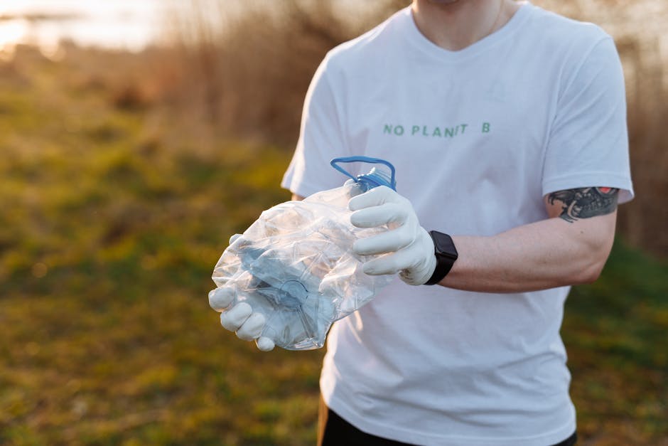 A pile of mixed waste materials is accumulated at the base of a rough concrete brick wall, situated outdoors on a dirt surface. The waste includes various cardboard boxes, some flattened and others partially expanded, with visible printed labels and tape, along with crumpled brown paper and plastic bags. A large, textured white bag made of woven fabric is partially covered by the waste, and a few small stones are scattered on the ground nearby. The scene is illuminated by natural daylight, creating soft shadows and highlighting the different textures of the cardboard, plastic, and fabric. The waste appears to be discarded materials awaiting collection or disposal, aligning with independent rubbish removal services such as those offered by Rubbish Removal Merton. The surrounding environment suggests an informal or back alley setting, with the waste's disorderly pile emphasizing the need for proper waste management or private clearance solutions to prevent clutter buildup and maintain cleanliness.