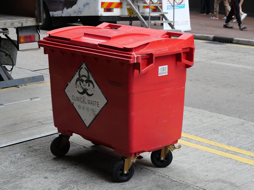 A large red plastic bin on four black caster wheels positioned on a paved urban street, used for disposing of clinical waste as indicated by a prominent white diamond-shaped hazard sign with black biohazard symbols and the label 'CLINICAL WASTE' in black text. The bin features a securely fitted lid with a slightly raised, contoured surface and a textured finish. Behind the bin, part of a truck with a grey and white design is visible, with metal stairs and signage for waste collection. The surrounding environment includes a section of sidewalk and a curb, with pedestrians walking along the street in the background, some wearing casual footwear. The scene is illuminated by natural daylight, with clear visibility of the bin's material, which appears durable and weather-resistant, and the overall setting suggests a designated area for waste collection and disposal, consistent with independent waste handling services.