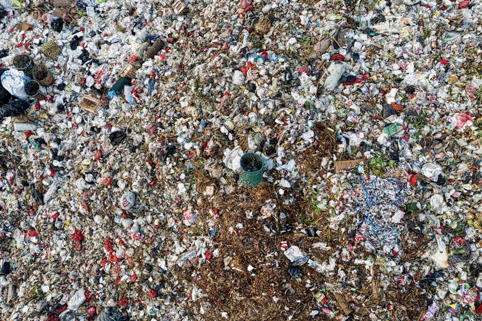 An expansive landfill site covered with a thick layer of mixed waste materials, including crumpled paper, plastic packaging, and discarded household items. Numerous plastic bags, some torn open, spill over the uneven surface of the refuse. Small pieces of cardboard, shredded cloth, and faded plastic bottles are scattered across the heap. In the middle of the image, a blue plastic barrel partially buried beneath the rubbish is visible among the clutter. The waste appears to be piled on a ground of compacted earth and debris, with some areas showing exposed dirt and organic matter. The surrounding environment looks exposed to natural light, highlighting the textures and various colours of the waste materials, creating a chaotic scene typical of unmanaged refuse deposits. The scene suggests the importance of professional rubbish removal services like those offered by Rubbish Removal Merton, emphasizing the need for alternative waste handling and on-site clearance solutions to manage waste efficiently and sustainably.