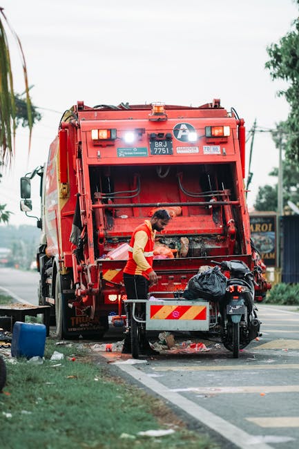 A large red plastic bin on four black caster wheels positioned on a paved urban street, used for disposing of clinical waste as indicated by a prominent white diamond-shaped hazard sign with black biohazard symbols and the label 'CLINICAL WASTE' in black text. The bin features a securely fitted lid with a slightly raised, contoured surface and a textured finish. Behind the bin, part of a truck with a grey and white design is visible, with metal stairs and signage for waste collection. The surrounding environment includes a section of sidewalk and a curb, with pedestrians walking along the street in the background, some wearing casual footwear. The scene is illuminated by natural daylight, with clear visibility of the bin's material, which appears durable and weather-resistant, and the overall setting suggests a designated area for waste collection and disposal, consistent with independent waste handling services.