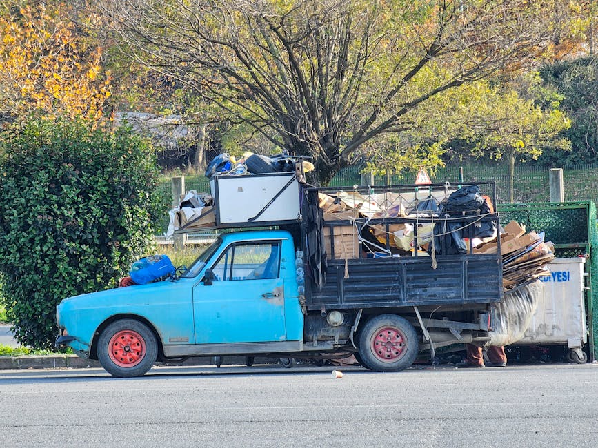 A small blue truck parked on the side of a street loaded with various types of rubbish and discarded items. The truck's front has a weathered, slightly faded blue paint finish, and the tires are a reddish-pink color. The cargo area is filled with a mixture of cardboard boxes, black garbage bags, wooden planks, and miscellaneous debris, some of which are loosely stacked and secured with ropes. In the background, there is a green park fence with vertical metal bars and a tall tree with light green and yellowish leaves, indicating a possible autumn setting. The environment appears to be a quiet residential area with soft natural daylight illuminating the scene. The scene reflects an alternative waste handling method, emphasizing the need for professional rubbish removal services like those provided by Rubbish Removal Merton for efficient on-site clearance of bulky items and waste.
