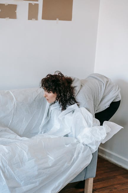 A woman with dark, curly hair, dressed in a light grey hoodie and dark trousers, is bending over on a grey upholstered sofa that is covered with white moving or packing sheets. She appears to be in the process of clearing or arranging items, with her head turned to the side and her hands out of sight. The setting is an indoor room with white walls, one of which has partially adhered or taped paper or cardboard pieces, possibly for decoration or renovation, visible in the background. The floor is made of dark wood, and natural light illuminates the scene, highlighting the textures of the sheets and furniture. This scene reflects activities associated with home decluttering, packing, or preparing for moving, which could involve the use of services like rubbish removal or waste management companies for disposing of debris or packing materials as part of an independent waste disposal process, typical of private or non-council collection options.