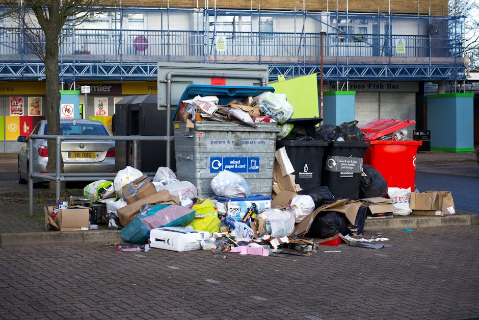 A large collection of mixed household and office waste is piled beside a metal recycling bin labeled for paper and cardboard. The pile includes flattened cardboard boxes, plastic bags filled with rubbish, crumpled paper, and various discarded packaging materials in different colors and textures. Some items, such as a white plastic container and a fabric bag, rest partially on the paved ground, while others, including black and black plastic trash bags, are stacked atop the bin and to its sides. The scene is set in an outdoor urban parking area near a commercial building with blue scaffolding and partially visible signage. A parked silver car is situated to the left of the rubbish pile, and a metal railing separates the waste area from the parking space. The environment appears to be a designated collection point for waste collection or private waste disposal, illustrating a typical scenario for rubbish removal services like those offered by Rubbish Removal Merton, in an area that requires alternative waste handling outside regular municipal collection.