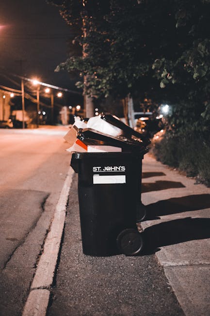 A black plastic rubbish bin with a slanted lid is positioned on a paved sidewalk along a quiet street at night. The bin is marked with the label 'ST. JOHN'S' and appears to contain various waste items, including paper and cardboard, some of which are protruding from the open lid. The surface of the bin has a matte finish, with some slight dust or dirt visible on it. The scene is illuminated by warm streetlights, creating soft shadows across the pavement and the side of the bin. In the background, there are blurred streetlights and the outlines of trees and parked vehicles, indicating an urban or suburban setting. The surrounding environment features a curb separating the sidewalk from the road, with some greenery on the right side. This image relates to the concept of rubbish collection and waste disposal, highlighting the importance of proper waste management, which companies like Rubbish Removal Merton facilitate through various waste handling services, including private waste collection outside of council provision.