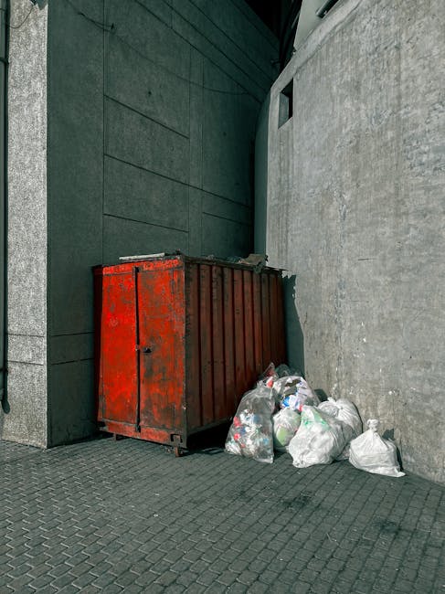 A red metal skip with a weathered, rusted surface is positioned against the corner where a dark green wall meets a light grey concrete wall in an urban outdoor setting. The skip's sliding door is closed, and it is situated on a paved surface composed of small, textured grey bricks. Next to the skip, on the right, are several white plastic bags filled with mixed waste, including visible plastic bottles and packaging. The bags rest on the pavement, some leaning slightly, and are gathered close to the base of the skip. The background features a narrow alleyway with the dark green wall extending vertically, and a small rectangular window high on the grey concrete wall, providing minimal natural light. The scene suggests a private, on-site waste collection related to rubbish removal services, possibly an alternative to council bin collections, with the skip and bags indicating recent waste disposal activity. Rubbish Removal Merton regularly handles such waste clearance, ensuring proper disposal of bulky refuse in urban environments.