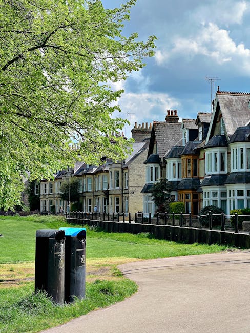 A row of traditional terraced houses with brick facades and bay windows, situated along a residential street. In the foreground, there are two black rubbish bins, some of which have visible scratches and weathering, placed on a grassy verge near a paved pathway. To the left, a large leafy tree with bright green foliage extends over the scene, providing shade and a natural element to the urban setting. The background features a partly cloudy sky with patches of blue, indicating a fair weather day. The scene is typical of a quiet suburban area where private waste collection might be arranged through local clearance services like Rubbish Removal Merton, which could facilitate on-site rubbish removal from such residential properties.