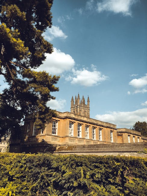 A historic stone building with classical architectural features, including large rectangular windows with white frames and decorative moldings, is visible in the image. The building's façade is made of beige-colored stone, textured with visible weathering and age, and it has a flat roof with a parapet. A tall, pointed spire tower, indicative of Gothic influence, rises from the center of the structure, featuring multiple pinnacles and intricate detailing. To the left, part of a large evergreen tree with dark green, dense foliage extends into the frame, casting shadows on the building’s exterior. The foreground consists of a neatly trimmed hedge and lush green grass, indicating a maintained garden or park setting. The scene is illuminated by natural daylight, with a bright blue sky dotted with a few fluffy white clouds overhead, suggesting a pleasant, clear day. This setting, captured by Rubbish Removal Merton, likely depicts a historic estate or civic building situated within a landscaped environment, relevant to alternative waste management or property clearance services in the Merton area.