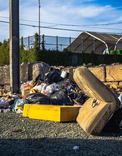 An outdoor scene shows a pile of mixed rubbish including black and clear plastic waste bags, a yellow plastic container, and discarded vehicle upholstery on a gravel surface. Behind the rubbish, there is a low stone wall composed of large, uneven stone blocks with rough textures, and a metal pole with electrical wires overhead. In the background, a hedge and a wire fence are visible, with a glimpse of a sports court or similar structure covered by a curved, semi-translucent roof, suggesting a recreational or community area nearby. The scene is bathed in natural daylight with a clear blue sky and some scattered clouds, indicating a bright, sunny day. This image illustrates an example of independent waste disposal, perhaps representing non-standard rubbish removal options that might be handled by private rubbish removal services like Rubbish Removal Merton, which manage bulky waste and general rubbish collection outside of local authority services.