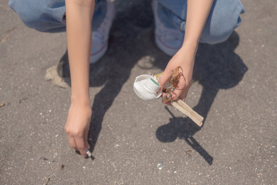 A close-up of a person's hand holding a small makeshift tool assembled from a wooden stick, a white plastic lid, and some natural fibers or roots, positioned above a paved concrete surface. The individual is wearing light blue jeans and white sneakers with red accents, and their arm and knee are partially visible in the background. The scene suggests an outdoor environment, possibly a sidewalk or driveway, with the person engaging in a cleanup or waste collection activity associated with rubbish removal services. The image emphasizes manual effort in handling small debris or refuse, highlighting the importance of individual participation in waste management and the potential for independent collection methods. The lighting is natural, casting a shadow of the hand and tool onto the ground, and the overall context aligns with themes of rubbish and waste handling, relevant to private rubbish removal options offered by companies like Rubbish Removal Merton.