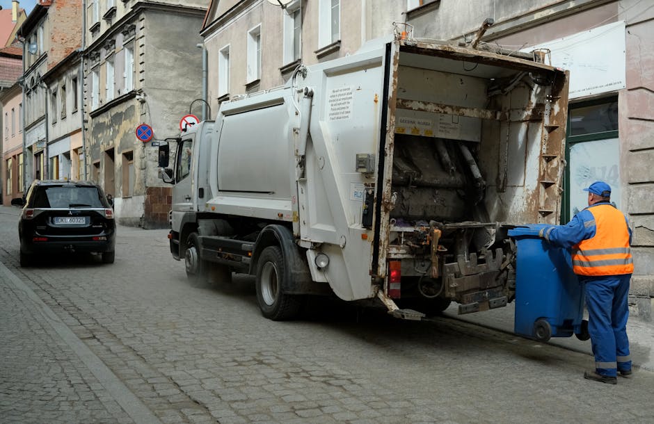 A sanitation worker wearing a blue uniform and an orange high-visibility vest is placing a blue wheeled refuse bin into the back of a white commercial rubbish collection vehicle on a city street. The truck, equipped with mechanical lifting and loading mechanisms, is parked adjacent to a row of older, multi-story buildings with stone and brick facades, some showing signs of wear and weathering. The pavement is paved with small, rectangular cobblestones, and a black car is parked further down the street. Two circular road signs are mounted on a pole near the buildings; one indicates no stopping and the other restricts certain vehicle actions. The scene is illuminated by natural daylight, creating a neutral, professional atmosphere that depicts routine urban rubbish collection, supporting the context of independent waste handling services in Merton.