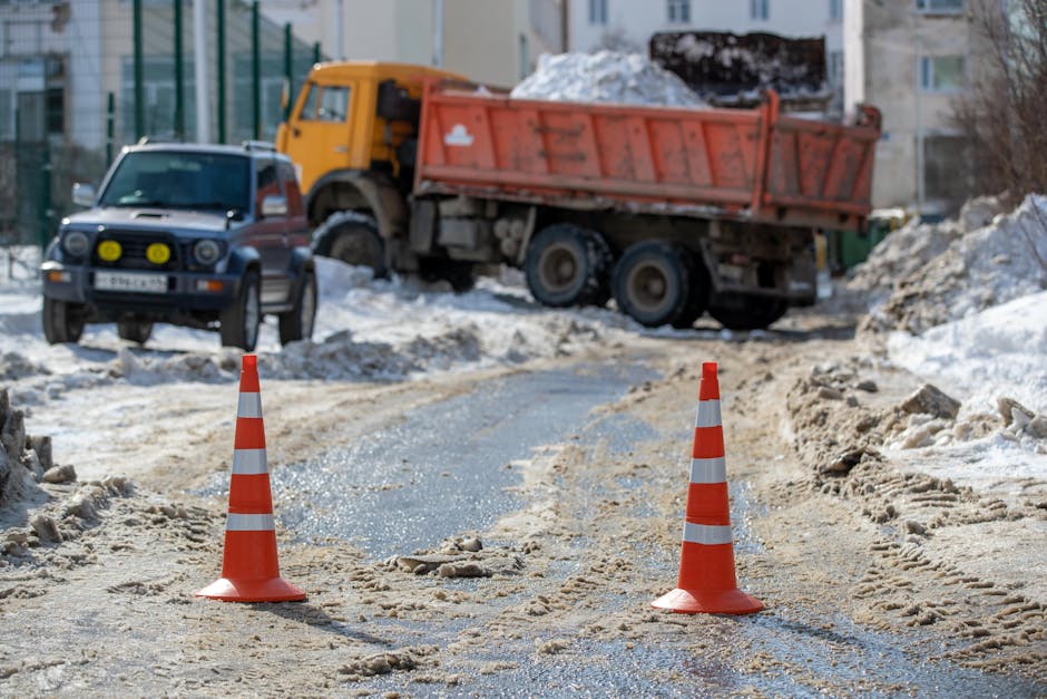 A construction scene on a snow-covered road features two orange and white striped traffic cones placed on either side of a partially cleared pathway. In the background, a large orange dump truck loaded with snow and debris is parked on the uneven surface, showing signs of dirt and wear on its exterior. To the left of the truck, a smaller dark-colored vehicle with yellow fog lights mounted on the front is situated on the roadway. The surrounding environment includes residential buildings and a fence, indicating an urban area where private or independent rubbish removal and snow clearance services might be operational. The scene is lit by natural daylight, emphasizing the texture of the snowy ground, the grit and mud on the road, and the weathered surfaces of the vehicles, highlighting the practical aspects of site clearance and waste handling in challenging winter conditions.