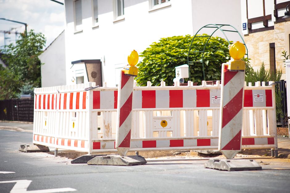 A construction site surrounded by a temporary white and red plastic barrier fence with reflective stripes, positioned on a paved street in a residential area. The barrier is equipped with yellow warning lights mounted on top of two vertical posts for visibility and safety. Behind the barrier, a large, well-manicured green shrub is visible, situated near a white building with several rectangular windows framed in dark brown. To the left, part of a black fence and some trees with green foliage can be seen, indicating a suburban environment. The scene is well-lit with natural daylight, highlighting the textures of the plastic barrier, the smooth asphalt, and the foliage. This setup suggests a private or alternative waste handling or site clearance activity, consistent with rubbish removal or on-site disposal services, such as those provided by Rubbish Removal Merton.