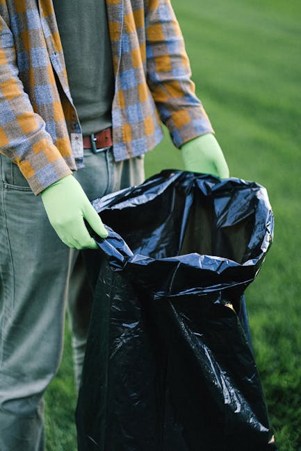 A person wearing green gloves, a checkered shirt in shades of blue, yellow, and brown, and a brown belt is seen holding a large black garbage bag open outdoors on a grass area. The bag appears to be made of plastic and is lined with a silver interior. The person is standing on what looks like a paved or grassy surface, and the background features a well-maintained green lawn with no other objects or structures visible. This scene is indicative of a rubbish collection activity, possibly an on-site clearance or private waste disposal, aligning with services offered by Rubbish Removal Merton. The natural lighting suggests daytime, and the person's attire indicates they are prepared for waste handling or environmental cleanup in an outdoor setting, supporting the context of rubbish removal or disposal support.