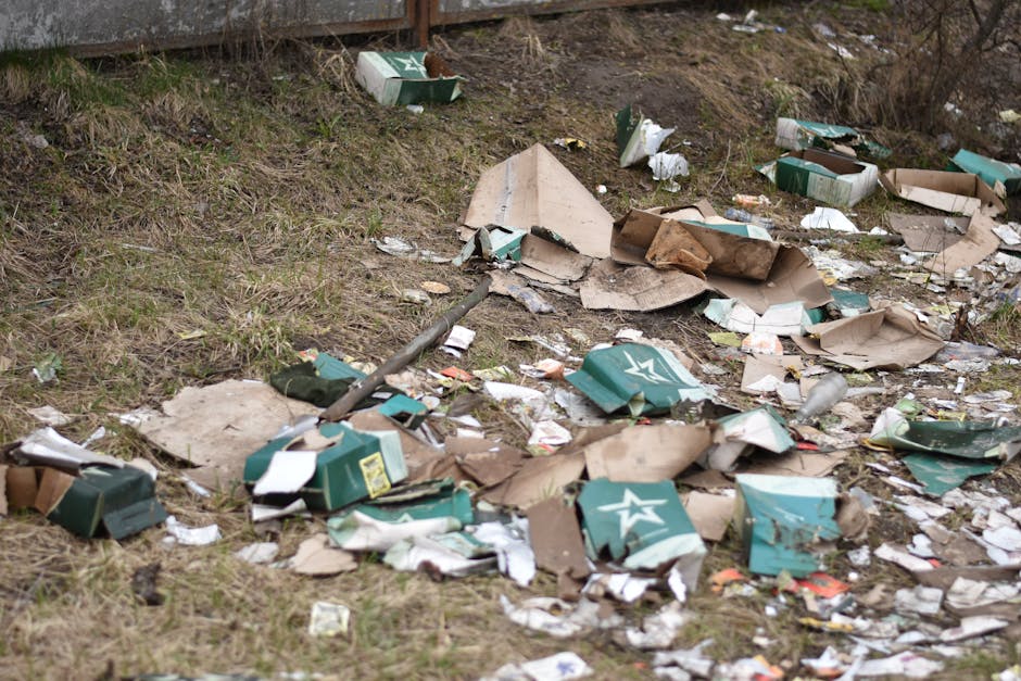 An outdoor scene showing a grassy area along a paved surface with scattered debris that includes torn cardboard boxes, some with green and white branding, and crumpled paper and plastic wrapping. A partially buried metal rod runs diagonally across the scene. The cardboard boxes are flattened and torn, with some pieces leaning against the grass, while others are spread across the ground. The environment appears to be a neglected space, possibly near a fence or retaining wall, with dirt and dried grass visible. The lighting suggests overcast or cloudy conditions, highlighting the disorganized state of the waste materials, which relate to private disposal or alternative rubbish removal options like on-site clearance, as managed by Rubbish Removal Merton, without any visible vehicles or additional equipment present.
